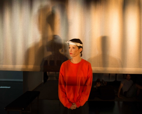 Zdjęcie ze spektaklu “Czarne papugi”. Kobieta w pomarańczowej bluzie i czapce z daszkiem stoi przed ekranem projektora. Na ekranie widać cienie innych postaci. Kobieta jest smutna.