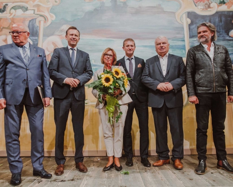 Na zdjęciu pięciu mężczyzn w garniturach i jedna kobieta stoją przed kurtyną wymalowaną według szkicu Stanisława Wyspiańskiego. Kobieta w dłoniach trzyma bukiet kwiatów.
