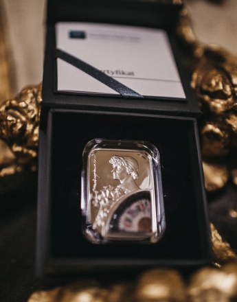 Na zdjęciu moneta z podobizną Heleny Modrzejewskiej. Srebrna, prostokątna w prawym dolnym rogu figury kolorowy wachlarz. Moneta umieszczona jest w granatowym futerale. Pudełko leży na fragmencie złotej rzeźby.
