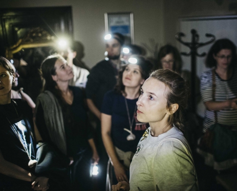 Na zdjęciu grupa ludzi stojąca w ciasnym pomieszczeniu. Niektórzy mają na sobie latarki czołówki założone na głowie. Wszyscy rozglądają się po pomieszczeniu. Na pierwszym planie stoi dziewczyna, wyglądająca na wystraszoną. Pomieszczenie jest ciemne.