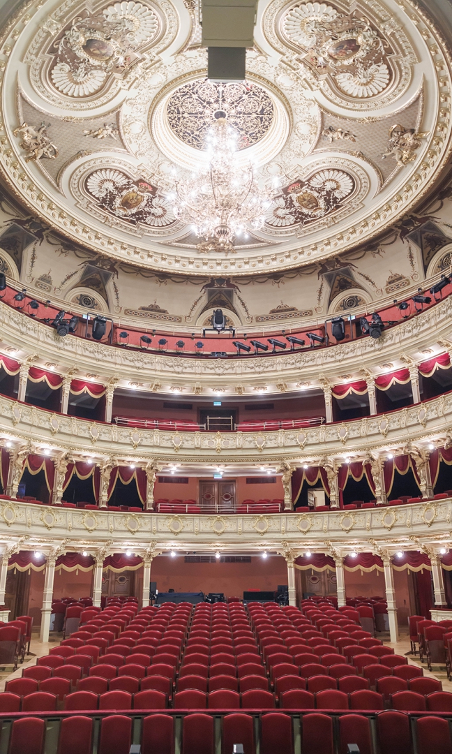 Zdjęcie pustej widowni teatru Słowackiego widzianej od strony sceny, Bordowe rzędy krzeseł, loże, bogato zdobione ściany w kolorze złota i ecru. Na trzecim piętrze na barierce zamontowane czarne reflektory. Na suficie ogromny żyrandol. Światła są rozświetlone, scena jest bardzo mocno oświetlona.