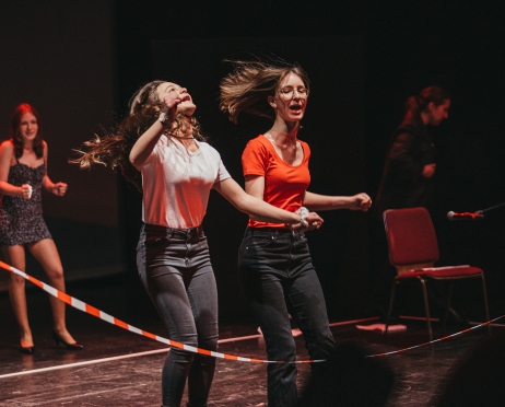 Na scenie grupa młodych kobiet. Scena oblana czerwonym światłem. Dwie kobiety przeskakują przez biało-czerwoną taśmę jak przez skakankę. Mają rozwiane długie włosy i otwarte usta. W tle tańczy kobieta w obcisłej mini i szpilkach.