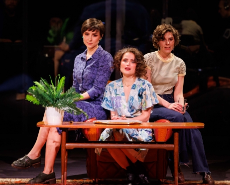 Na zdjęciu w centralnej części na środku trzy kobiety. Siedzą przy ławie w kolorze brązowym. Są smutne. Patrzą prosto przed siebie. Kobieta po lewej stonie ubrana jest w niebieska sukienkę, obok niej kobieta ubrana w niebieską sukienkę w kolorowe kwiatki. Po prawej stronie kobiet ubrana w bluzkę w kratkę. Na ławie zielony kwiatek. Zdjęcie jest minimalistyczne. ło jest czarne.