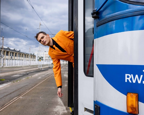 Wagon tramwajowy w kolorze niebiesko-białym sfotografowany z czoła wagonu. Wagon ma otwarte drzwi z których wychyla się do połowy pasa młody mężczyzna w pomarańczowym garniturze. W tle budynki i zachmurzone niebo.