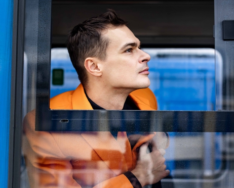 W oknie tramwaju siedzi skulony mężczyzna w pomarańczowym garniturze. Ma odsuniętą szybę w oknie, patrzy w bok- twarz jest widoczna z profilu. Wygląda na zamyślonego.