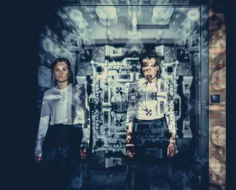 Na zdjęciu dwie dziewczyny stojące obok siebie. Obie ubrane są w białe koszule i czarne spódnice. Stoją pod ścianą. Na nich wyświetlana jest projekcja drobnych elementów, przypominających wtyczki i przyciski.