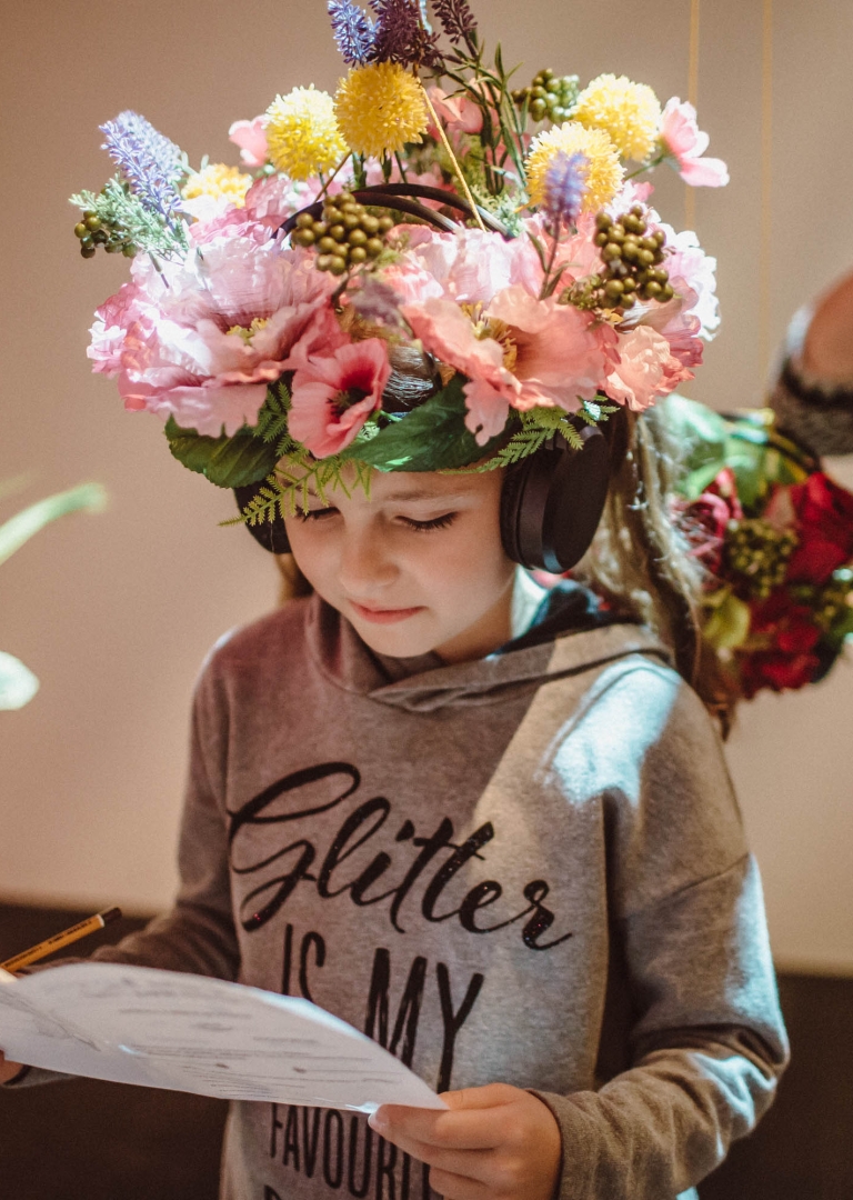 Dziewczynka w wianku ze sztucznych kwiatów czyta z kartki trzymanej w dłoniach. Na uszach ma duże czarne słuchawki audio, w dłoni ołówek. W wianku dominują różowe i żółte kwiaty. W lewym rogu zdjęcia wisi podobny wianek. Za dziewczyną stoi uśmiechnięta kobieta z takimi samymi słuchawkami na uszach.