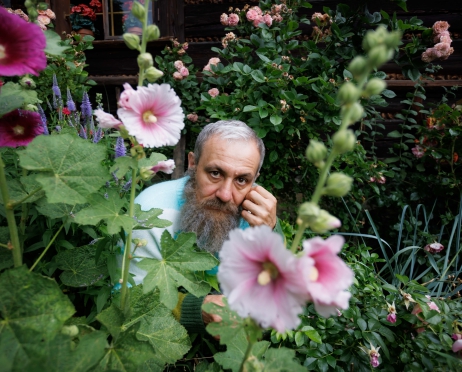 Ogród pełen zieleni i wysokich kwiatów. Zza różowych malw wychyla się starszy mężczyzna, z brodą wokół twarzy, w błękitnym sweterku. Ma brązowe oczy, patrzy w obiektyw, dotyka dłonią twarzy.