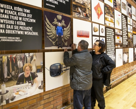 Zdjęcie zrobione w Domu Rzemiosł Teatralnych. Na zdjęciu kobieta i mężczyzna. Stoją przy ścianie ze zdjęciami. Zdjęcie zrobione z boku. Kobieta patrzy na zdjęcia mężczyzna coś dorysowywać na jednym ze zdjęć. Mężczyzna stoi tyłem, kobieta bokiem. Ręką podpiera się na biodrze.