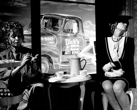 Czarno-białe zdjęcie przedstawia dwie kobiety siedzące przy małym, okrągłym stoliczku. Pierwsza z nich ubrana w luźną sukienkę. Patrzy na swoje paznokcie. Druga kobieta ubrana w elegancką sukienkę i kaszkiet. Na dłoniach ma rękawiczki. Odwraca głowę. Na stoliku dwie filiżanki i dzbanek. W tle samochód pick-up.