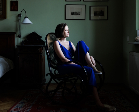 Na zdjęciu kobieta w niebieskim spodnium siedzi na krześle bujanym. Jedną nogę trzyma na siedzisku krzesła. Patrzy w okno znajdujące się przed nią. Za nią łóżko z ciemnobrązowym drewnianym zagłówkiem. W tle zielona ściana na której znajdują się rysunki.
