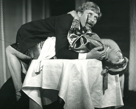Czarno-białe zdjęcie ze spektaklu “W małym dworku”. Mężczyzna i kobieta leżą na stole zmysłowo się obejmując. Kobieta ma na sobie ozdobną sukienkę a mężczyzna garnitur oraz szkiełko w oku.