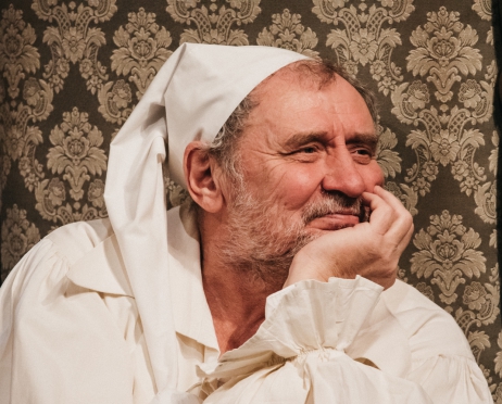 Na zdjęciu aktor Andrzej Grabowski, zdjęcie od klatki piersiowej w górę. Aktor ubrany w białą koszulę i białą szlachmycę na głowie. uśmiecha się, podpiera podbródek ręką, patrzy w dal. Siedzi na krześle z wysokim oparciem z obiciem w brązowe ornamenty