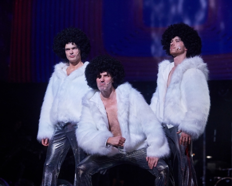 Na zdjęciu trzech mężczyzn ubranych w białe krótkie futra i srebrne, błyszczące spodnie. Mężczyźni mają na głowie jednakowe czarne afroperuki i zacięte wyrazy twarzy. Mężczyzna centralnie lekko przysiadł na ugiętych kolanach. Pozostali stoją