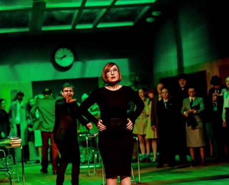 Zdjęcie ze spektaklu “Wyzwolenie”. Na pierwszym planie kobieta w czarnej sukience, za nią mężczyzna. Wydaje się. że próbuje ją zawołać. Scenografia przypomina klasę szkolną. Scena oświetlona jest zielonym światłem.