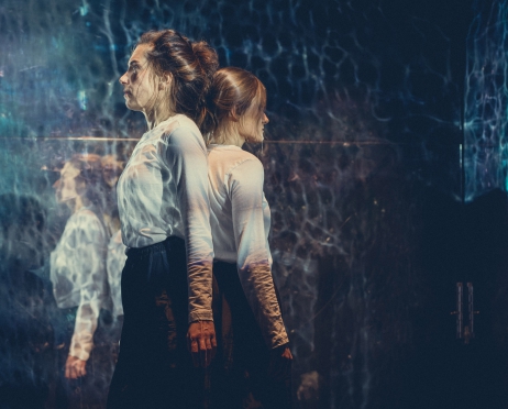 Dwie dziewczyny stoją plecami do siebie. Ubrane są w takie same białe bluzki i czarne spódnice. Patrzą przed siebie. Na ścianie za nimi wyświetlana jest projekcja przypominająca wodę, w której odbija się światło.