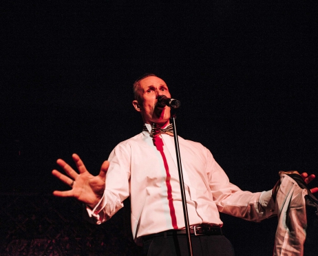 Zdjęcie ze spektaklu “Minia-tour. Muzyczne biuro podróży”. Na zdjęciu mężczyzna śpiewa do stojącego mikrofonu. ma na sobie białą koszulę i muszkę. Tło jest czarne.