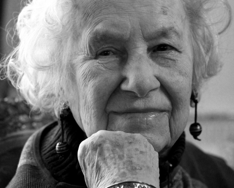 Czarno-biały portret kobiety. Staruszka ma siwe włosy, podpiera głowę dłonią. Spogląda przed siebie. Jej twarz pokryta jest zmarszczkami. Jest radosna.