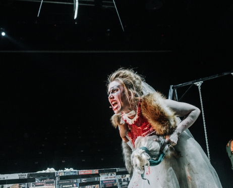 Zdjęcie ze spektaklu “Bolesław Śmiały”. Na zdjęciu kobieta w białej sukience, poplamiona krwią, krzyczy. Za nią znajduje się metalowa huśtawka. Obok niej stoją poukładane kasety VHS. Tło jest czarne.