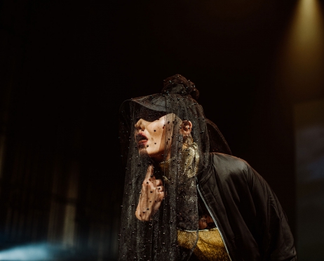 Zdjęcie ze spektaklu “Wyspiański. Koncert”. Kobieta na zdjęciu ubrana jest w czarną kurtkę i czarny welon narzucony na twarz. Tło jest czarne.