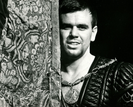 Czarno-biała fotografia ze spektaklu “Hamlet”. Portret młodego mężczyzny wychylającego się zza kotary. Mężczyzna ma na sobie skórzany kubrak i medalion na szyi.