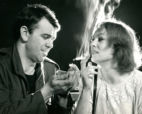 Czarno-białe zdjęcie ze spektaklu “Ławeczka”. Młody mężczyzna zapałkami przypala papierosa siedzącej obok niego kobiecie. Kobieta spogląda na mężczyznę. Całą scenę spowił papierosowy dym.