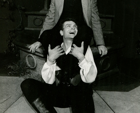 Czarno-biała fotografia przedstawia dwóch mężczyzn. Jeden z nich siedzi na podłodze. Drugi mężczyzna siedzi na podwyższeniu zaraz za nim i obejmuje go nogami. Obaj są bardzo radośni, śmieją się do siebie.