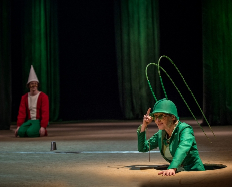 Zdjęcie ze spektaklu “Czarnoksiężnik z Krainy Oz”. Na pierwszym planie znajduje się kobieta w zielonym kostiumie z czapką przypominającym konika polnego. Wydaje się, że wychodzi spod ziemi. Na drugim planie znajduje się chłopak w kostiumie Pinokia, ubrany w spiczasty kapelusz i długi nos. Tło jest zielone.