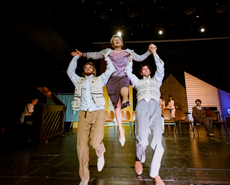 Zdjęcie ze spektaklu “Turnus mija a ja niczyja. Operetka sanatoryjna”. Fotografia przedstawia dwóch mężczyzn, którzy podnoszą radosną kobietę. Ubrania tych osób przypominają modę czasów PRL. Z tyłu  po lewej stronie znajduje się dziewczyna grająca na pianinie. W tle ustawione są krzesła oraz kolorowe elementy scenografii.