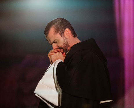 Zdjęcie ze spektaklu “Imię róży”. Mężczyzna na zdjęciu jest ustawiony bokiem, składa ręce do modlitwy. Włosy ma na gładko zaczesane do tyłu. Jego ubranie przypomina czarną szatę mnicha. Tło jest czarne.
