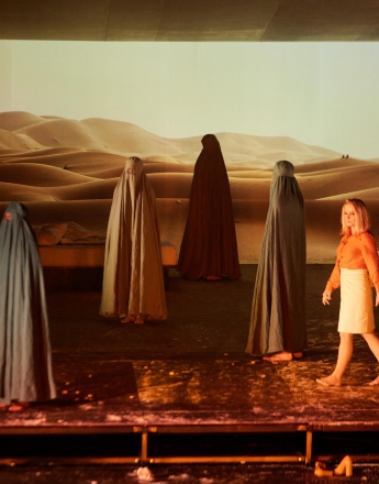 Na zdjęciu kobieta w jasnej spódnicy i brązowej koszuli przechodzi przez scenę. Wokół niej stoi sześć postaci w muzułmańskim stroju zakrywającym całe ciało, włosy i oczy. W tle wyświetlane jest zdjęcie pustyni. Podłoga jest złota. Panuje półmrok.