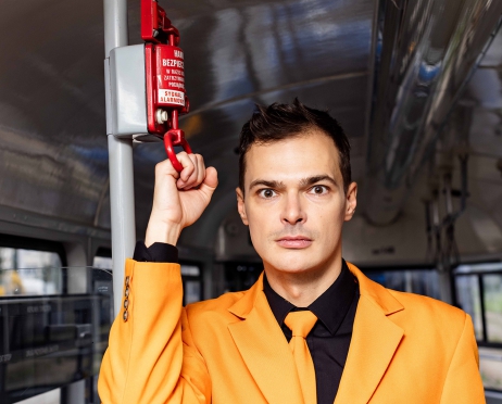 Zdjęcie mężczyzny w pomarańczowym garniturze i czarnej koszuli, widoczny od pasa w górę. Stoi w pustym wagonie tramwajowym, trzyma dłoń na zwisającym z góry czerwonym hamulcu bezpieczeństwa. Ma zdziwioną minę, wpatruje się w obiektyw.