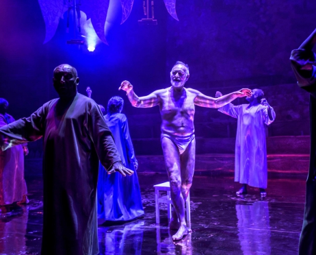 Na zdjęciu grupa postaci tańczących w długich szatach W centralnym miejscu stoi starzec w samych slipach, ma uniesione ręce do góry. Na zdjęciu dominuje ciemne, niebieskie i fioletowe światło.