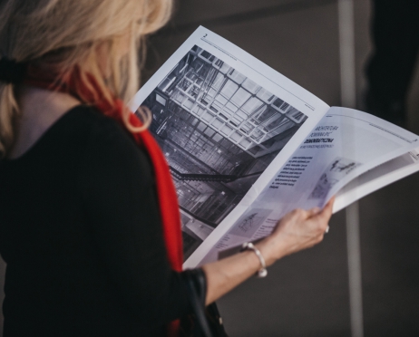 Zdjęcie przedstawia wnętrze Małopolskiego Ogrodu Sztuki. Zdjęcie zrobione z góry. Na zdjęciu kobieta czyta gazetę. Zajmuje całą powierzchnię zdjęcia. Ubrana jest w czarną sukienkę i czerwony szal. Tło jest szare.