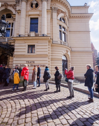Na zdjęciu grupa ludzi stoi w kolejce przed wejściem do budynku. Na elewacji jest napis Teatr im. Juliusza Słowackiego w Krakowie.  Przed budynkiem jest brukowany chodnik.