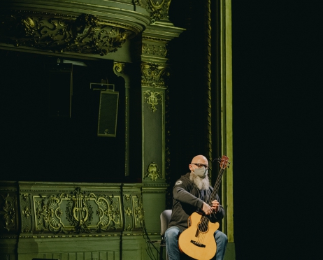 Mężczyzna siedzi na krześle i trzyma gitarę. Jego twarz jest zasłonięta materiałową maską. W tle fragment widowni Dużej Sceny Teatru Słowackiego. Widownia jest ciemna. Światło pada tylko na mężczyznę.