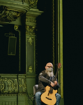 Mężczyzna siedzi na krześle i trzyma gitarę. Jego twarz jest zasłonięta materiałową maską. W tle fragment widowni Dużej Sceny Teatru Słowackiego. Widownia jest ciemna. Światło pada tylko na mężczyznę.