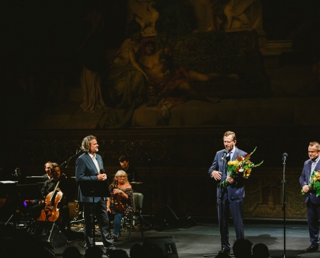 Na zdjęciu scena. Po prawej stronie dwóch mężczyzn. Marszałek oraz Wiceprezydent Krakowa. Trzymają kwiaty. Jeden z mężczyzn przemawia. Po lewej stronie bikiem sti dyrektor Teatru. patrzy w ich stronę. Stoi bokiem. Za Mężczyzną po lewej stronie orkiestra. W tle kurtyna Siemiradzkiego.
