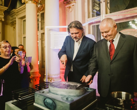 Zdjęcie zrobione we foyer Teatru. W centralnej części praktycznie na całej powierzchni dyrektor Teatru i prezes NBP. Kroją tort. Są uśmiechnięci. Po lewej stronie stoi kobieta robi im zdjęcie. Tort jest w kształcie monety. W kolorze szarym.