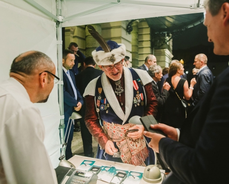 Zdjęcie zrobione przed budynkiem teatru z namiotu. Mężczyzna z bractwa kurkowego stoi nad stolikiem. Ogląda monety. Na pierwszym planie dwóch mężczyzn stoją bokiem. Mówią do niego, patrzą w jego kierunku. W lte grupki osób. Rozmawiają ze sobą. W tle budynek Teatru.