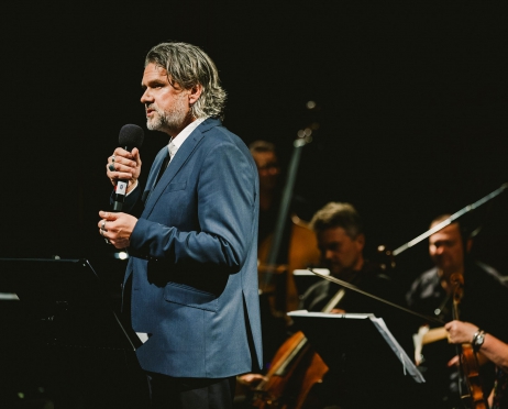 Na zdjęciu po lewej stronie na pierwszym planie tułów dyrektora Teatru. Stoi bokiem. przemawia. Jest skupiony. Ubrany jest w granatowy garnitur. W tle orkiestra. Tło jest czarne.