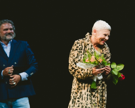 Na zdjęciu w centralnej części aktorka Hanna Bieluszko. stoi lekko bokiem trzyma kwiaty patrzy w prawą stronę. Uśmiecha się. Po lewej stronie dyrektor teatru. W ręku trzyma mikrofon. Patrzy w stronę kobiety. Uśmiecha się. Tło jest czarne.