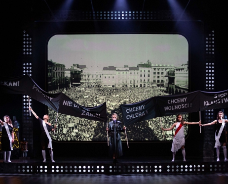 Na zdjęciu pięć kobiet stoi na pustej scenie. W dłoniach trzymają czarne transparenty z białymi napisami: Chcemy chleba, chcemy wolności, stop kłamstwom. W tle wyświetlone czarno-białe zdjęcie tłumu ludzi zgromadzonych na płycie krakowskiego rynku.