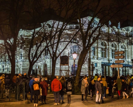 Zdjęcie zrobione z Plant. Na pierwszym planie grupa ludzi stojąca tyłem. Patrzą w stronę budynku teatru który jest oświetlony białym światłem. zaznaczone są ciemniejszym światłem kontury budynku. Budynek przebija się przez drzewa bez liści. Tło jest czarne.