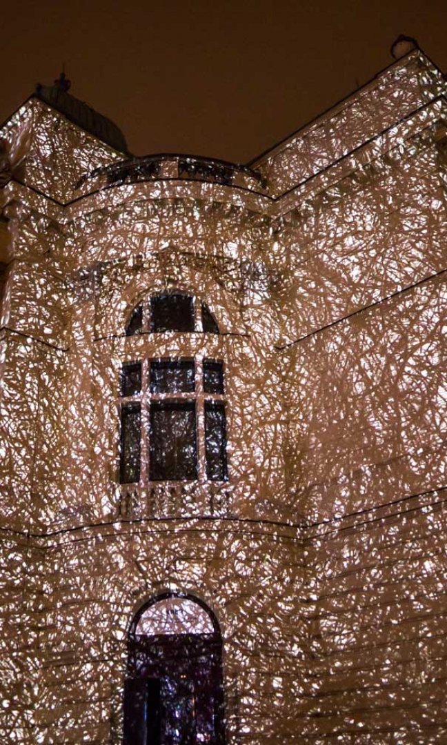 Zdjęcie nocą. Na zdjęciu fragment półokrągłej fasady budynku z oknami. Na budynku wyświetlona projekcja przypominająca poplątane suche konary drzew.