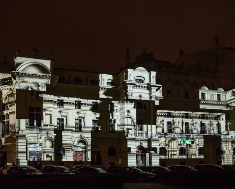 Na zdjęciu fragment budynku głównego Teatru od strony parkingu. Kanciasta budowla oświetlona na biało z kwadratowymi mniejszymi ciemniejszymi figurami. Zdjęcie jest tajemnicze. Tło jest czarne.