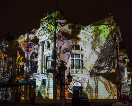 Na zdjęciu w centralnej części na całej powierzchni fragment głównego budynku Teatru. Fasada jest oświetlona. Kolorowe światła przedstawiają twarz Wyspiańskiego. Twarz jest rozłożona na całym fragmencie fasady. Tło jest czarne.