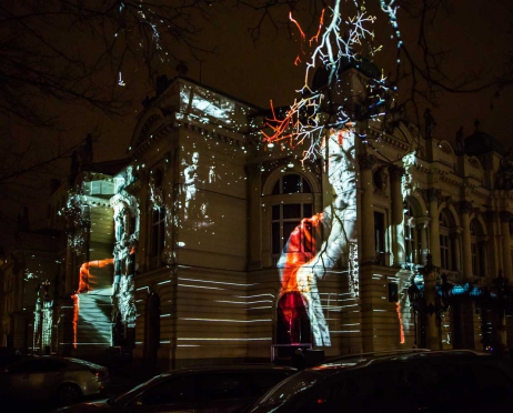 Zdjęcie zrobione z boku. Frontowa część budynku Teatru Słowackiego oświetlona w postaci ze spektaklu “Imię róży” Kolory biało czerwone. Fragment budynku znajduje się na całej powierzchni zdjęcia w centralnej części. Tło jest czarne.