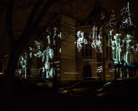 Na zdjęciu w centralnej części na całej powierzchni fragment głównego budynku Teatru. Fasada jest oświetlona na biało. Mapping przedstawia aktorów z różnych spektakli granych w Teatrze,. Zdjęcie jest tajemnicze. Tło czarne.