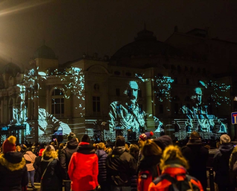 Zdjęcie zrobione od strony ul. Szpitalnej. Na pierwszym planie grupa ludzi stojąca tyłem. Patrzą w kierunku budynku głównego Teatru. Budynek świeci się na biało, wyświetla się na nim postać pojawiająca się na fasadzie kilka razy, cyklicznie. Tło jest czarne.